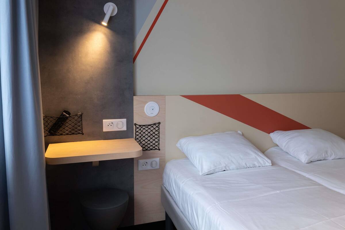 A modern hotel room with a bed, bedside table, and a wall-mounted light fixture.