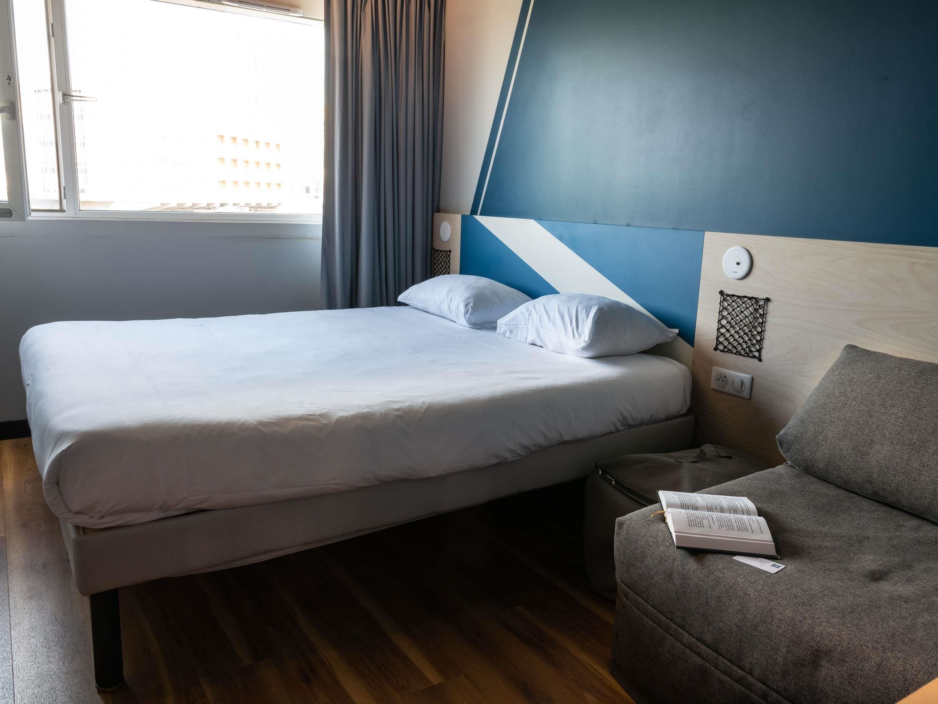 A hotel room with a bed, a grey sofa, a window with curtains, and a wall with a geometric design.