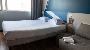 A hotel room with a bed, a grey sofa, a window with curtains, and a wall with a geometric design.