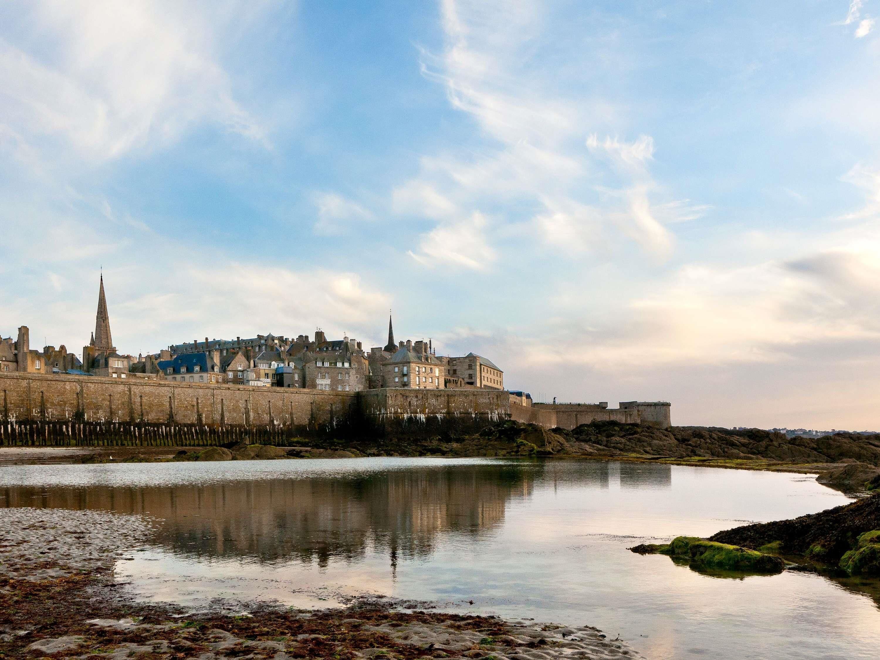 A coastal cityscape with historic buildings, a prominent tower, and a calm body of water reflecting the structures.
