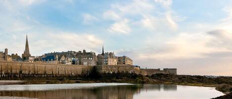 A coastal cityscape with historic buildings, a prominent tower, and a calm body of water reflecting the structures.