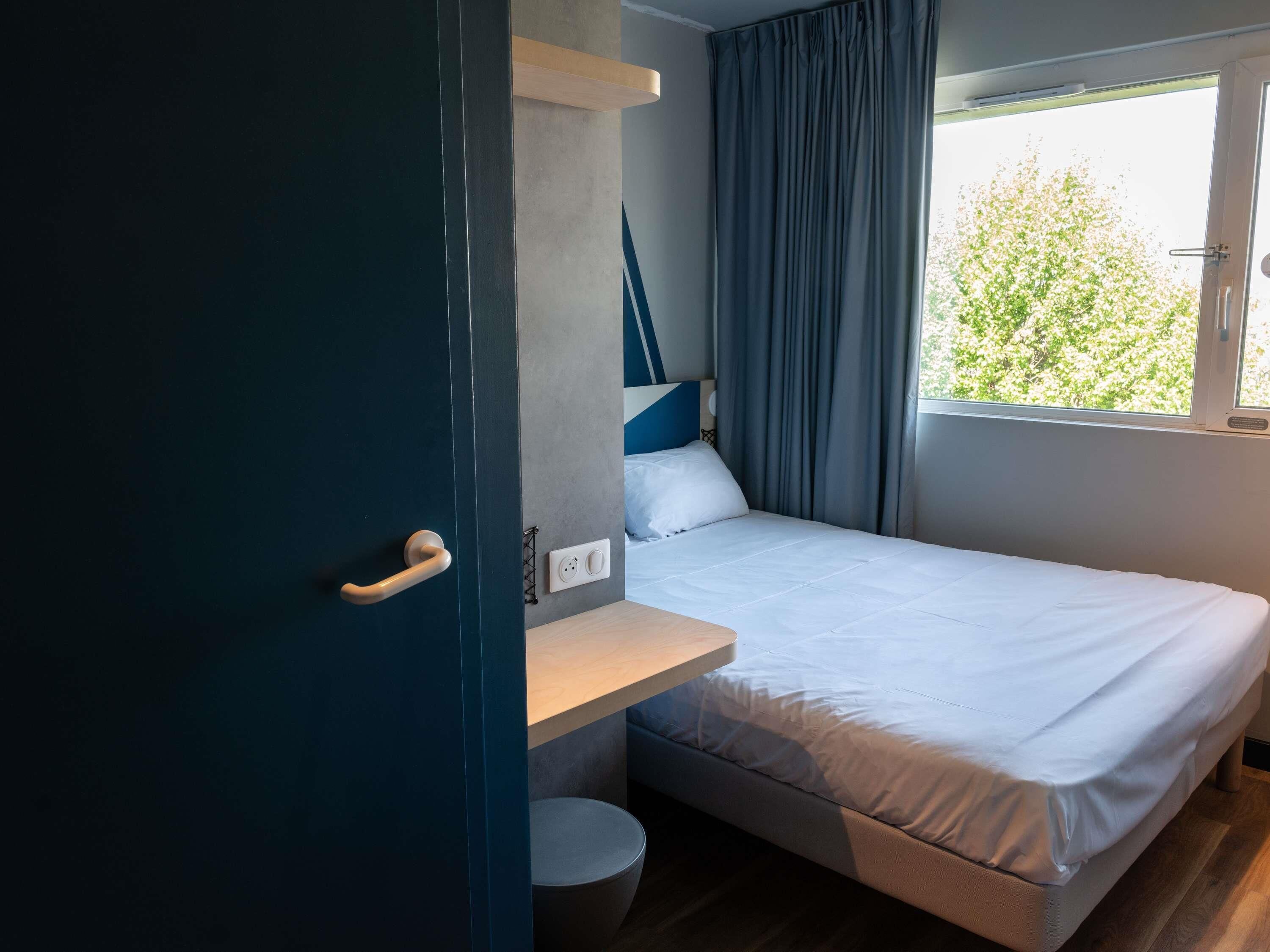 Habitació petita i funcional amb un llit individual, una taula de nit reduïda, una finestra amb cortines i una porta.