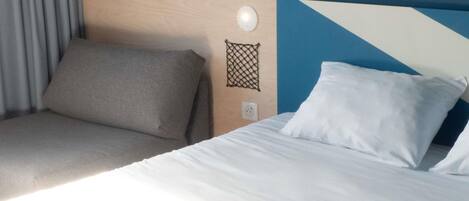 Quarto de hotel com cama, cabeceira, luminária fixada na parede e tomada.