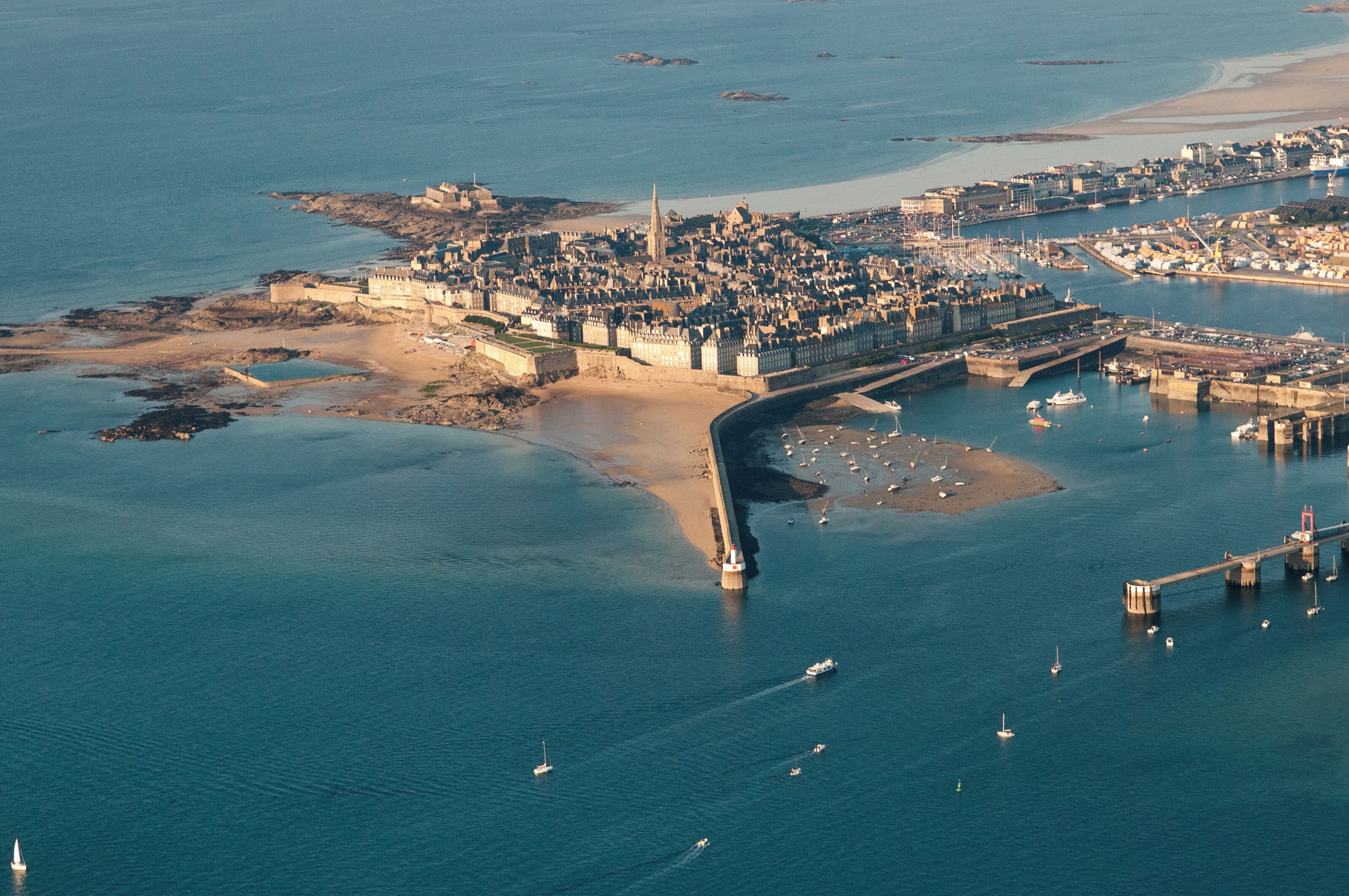 An aerial view of a coastal city with a prominent castle, a lighthouse, and a harbor with boats.