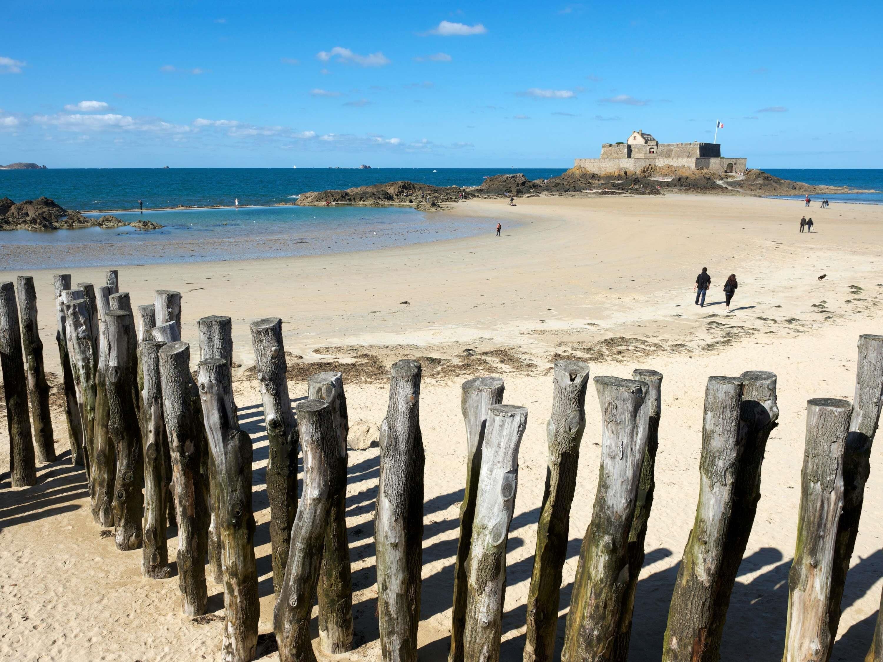Een zandstrand met een houten omheining op de voorgrond, een rotsachtige richel, en een gebouw op een heuvel in de verte.
