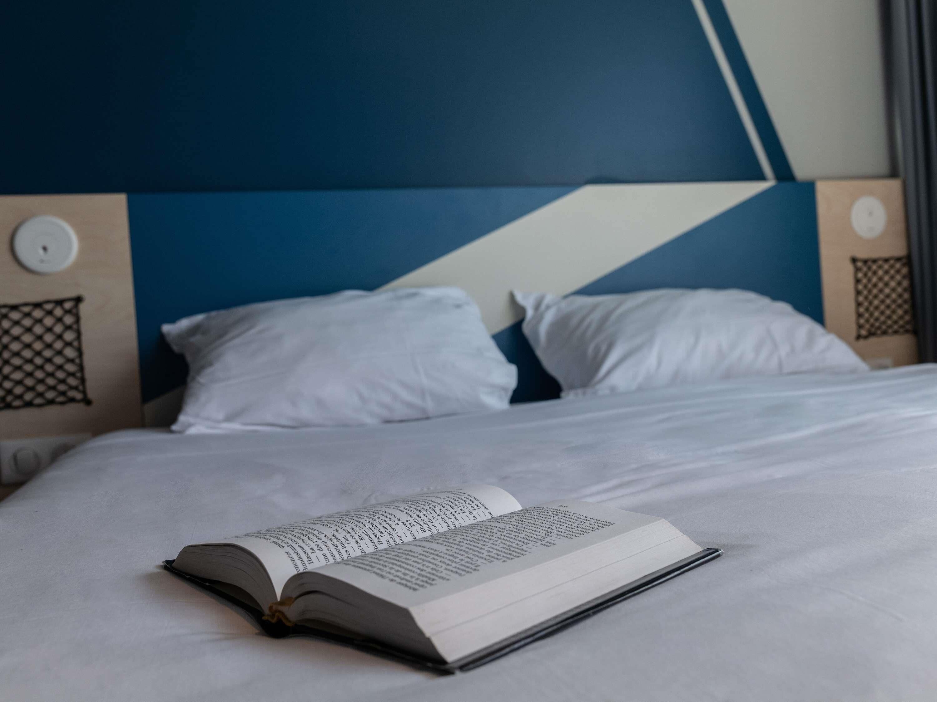A neatly made bed with white bedding and pillows, a book open on the bed, and a modern headboard with geometric patterns.