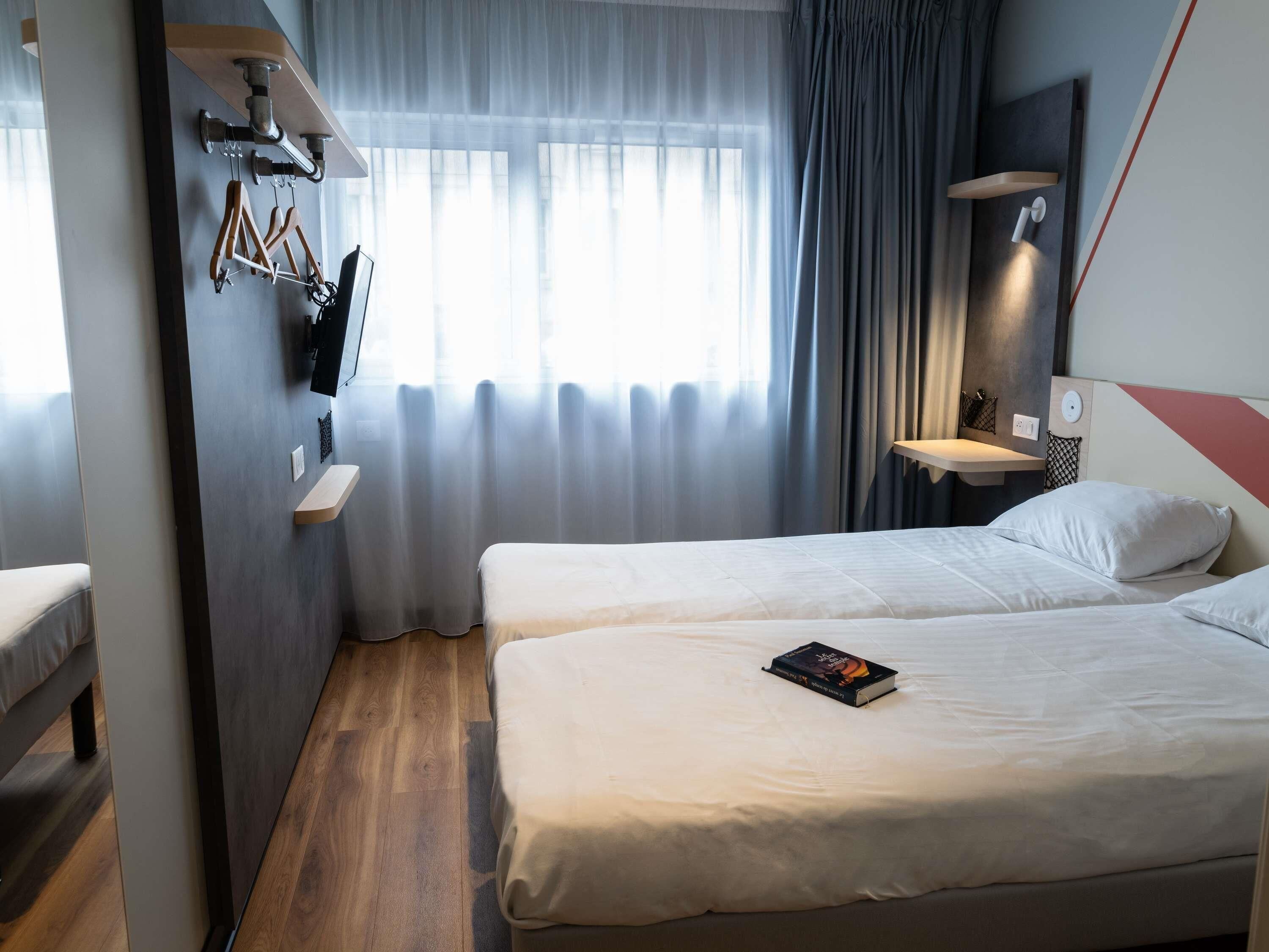 A hotel room with two beds, a wooden floor, a window with sheer curtains, and a wall-mounted shelf with a TV and clothes.