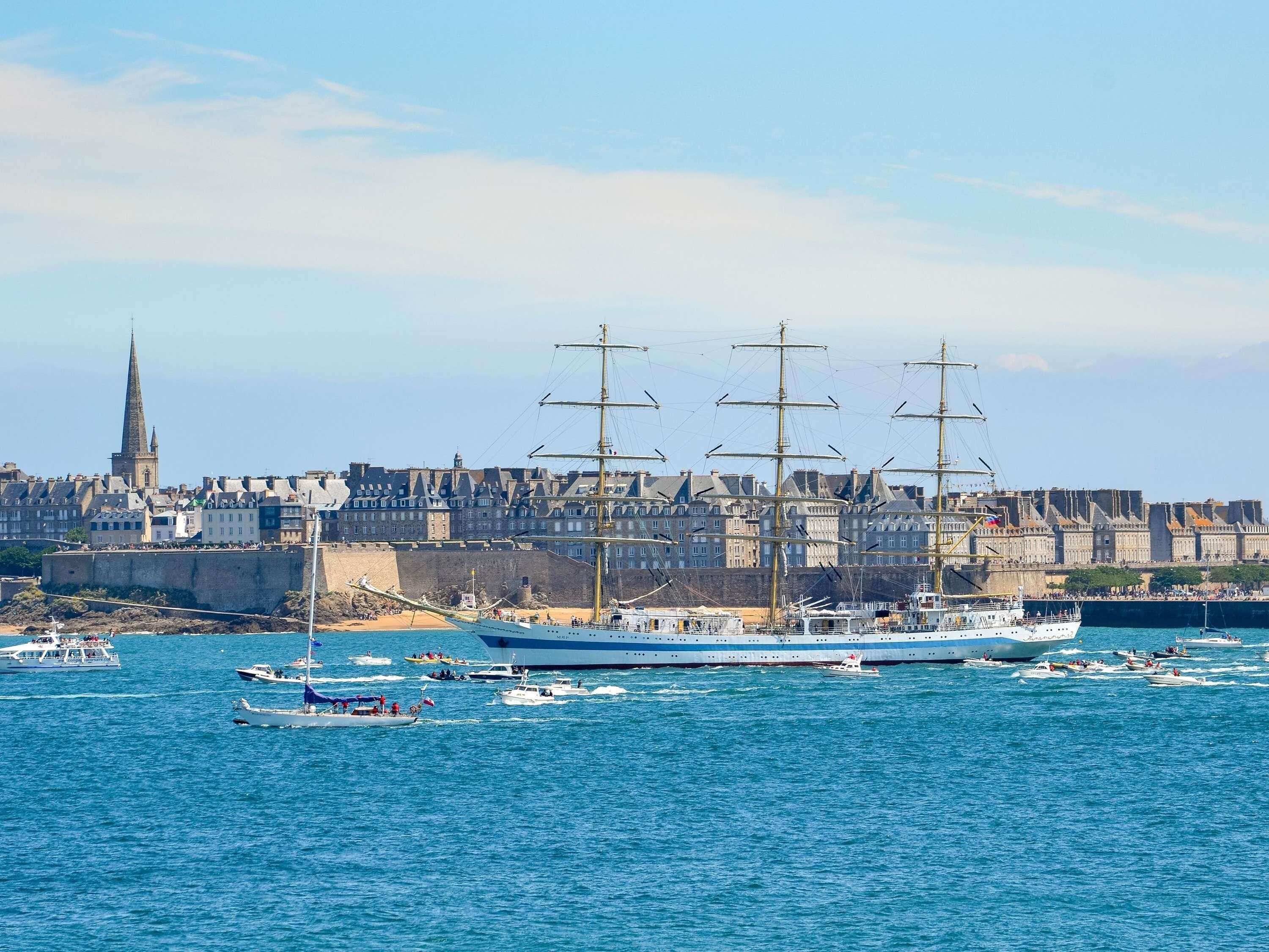 Un puerto con varios barcos, un buque grande y un paisaje urbano con edificios históricos al fondo.