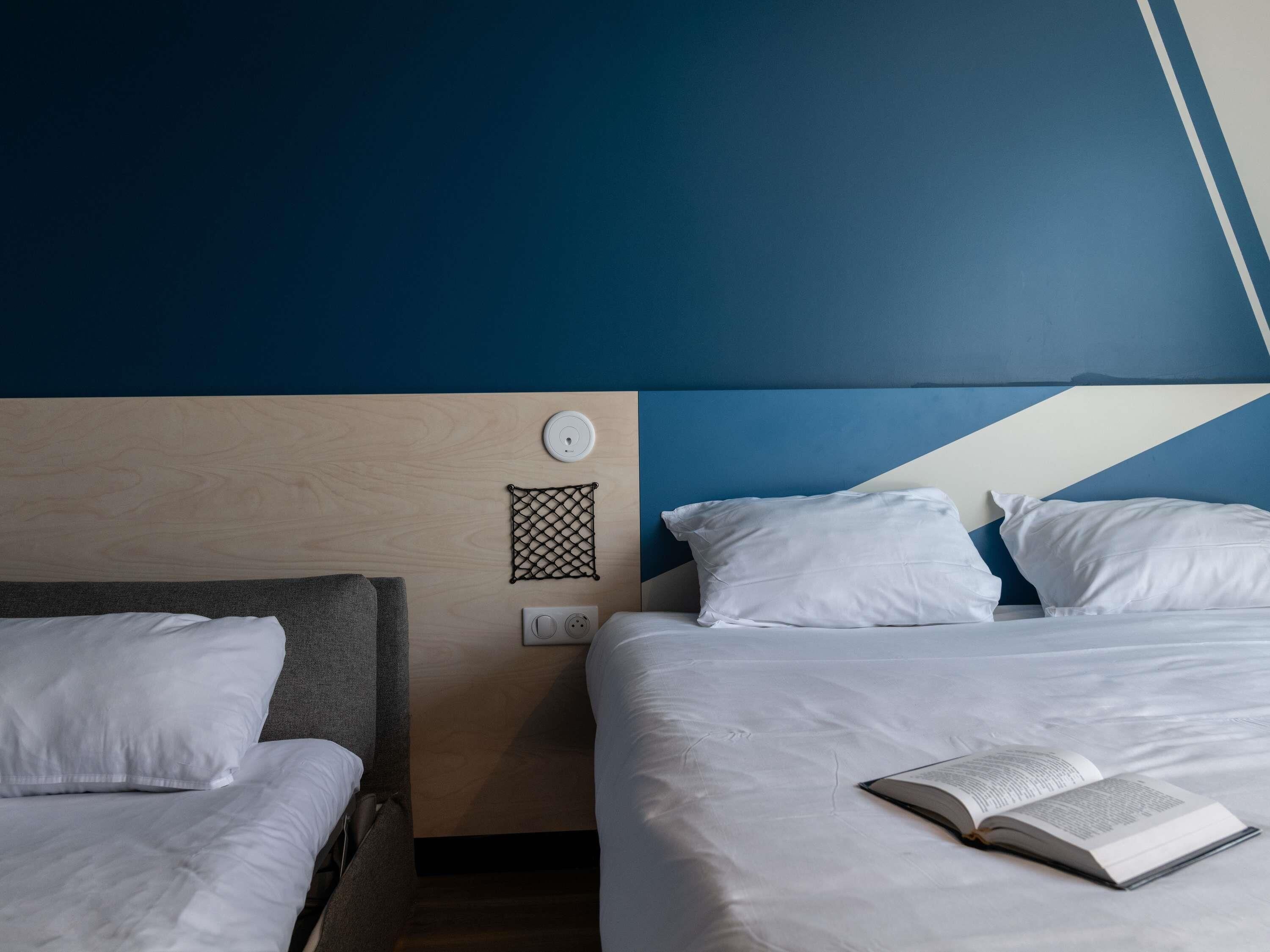 Habitación de hotel con dos camas, cabeceros de madera, un panel de control en la pared y un libro sobre la cama de la derecha.
