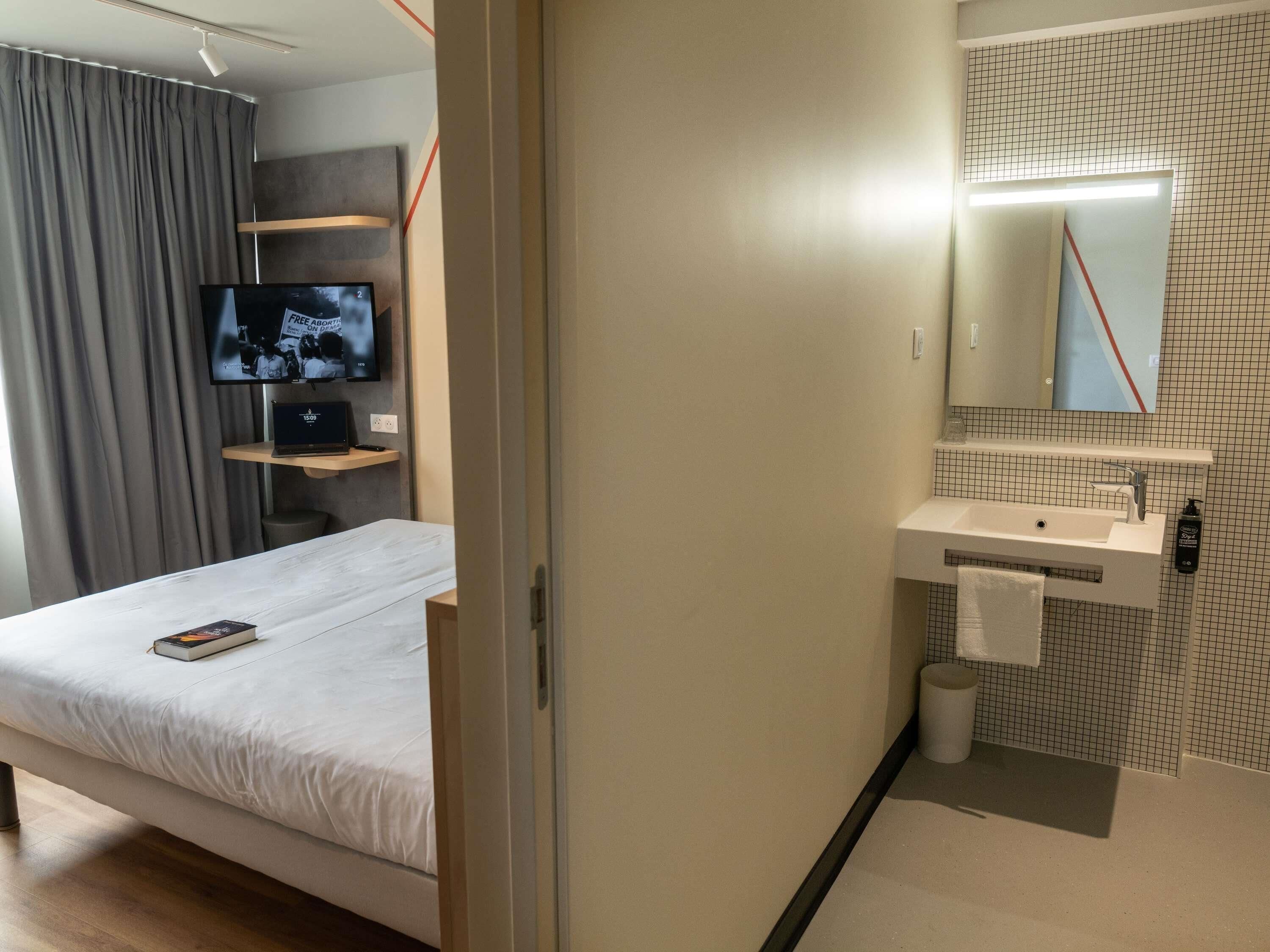 Une chambre d’hôtel comprenant un lit, une télévision, un bureau, une salle de bain avec un lavabo et un miroir, et une douche.