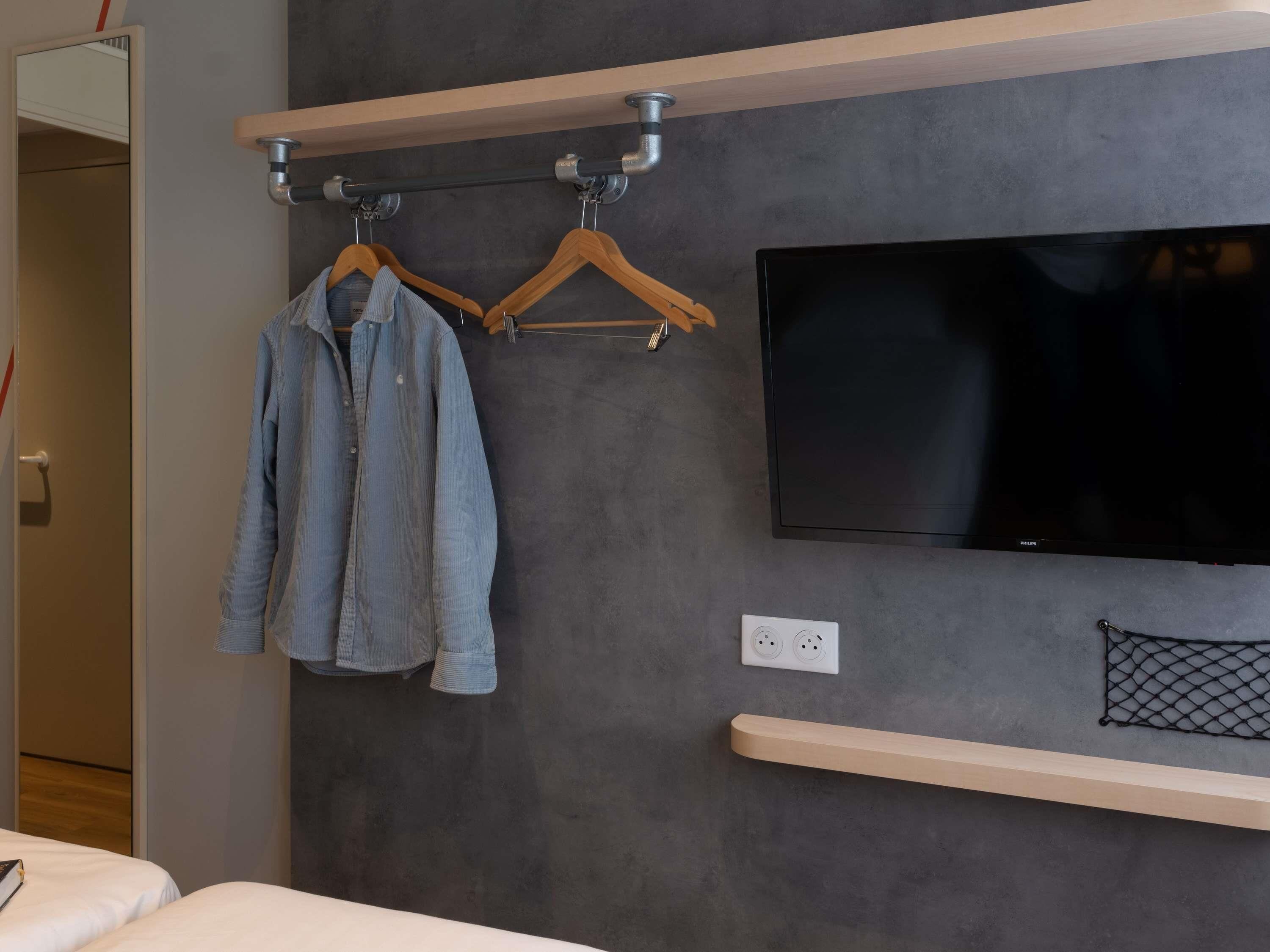 A modern hotel room with a flat-screen TV mounted on the wall, a wooden shelf below, and a clothing rack with a denim shirt.