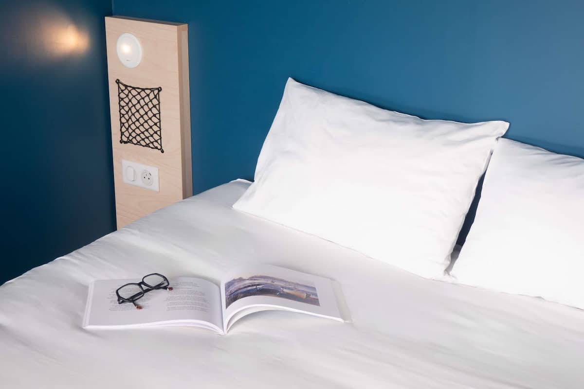 A neatly made bed with white linens, a pair of glasses, and an open book on the bed.