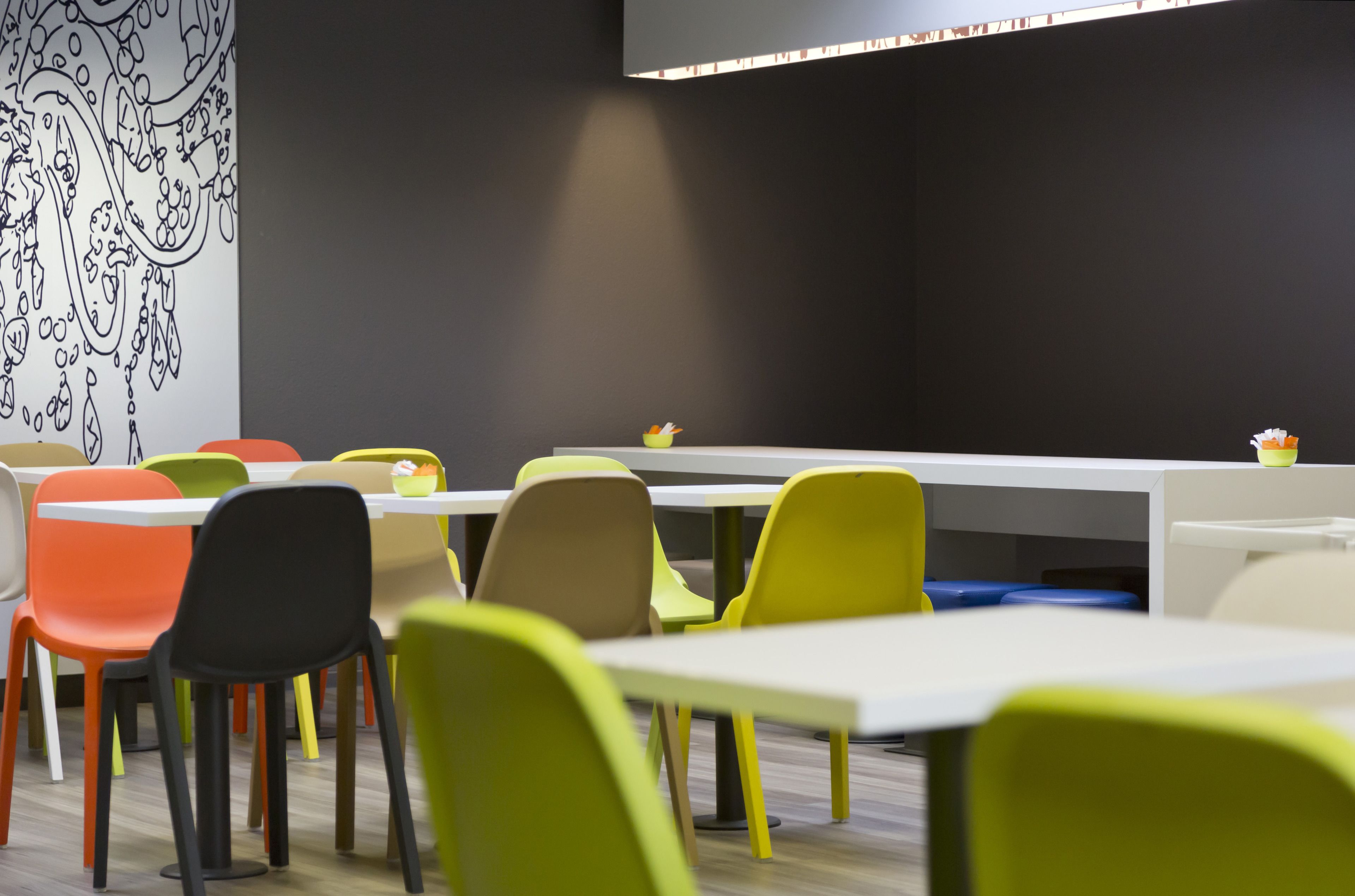 Un espace repas moderne avec des chaises et des tables aux couleurs vives, une fresque murale en noir et blanc, et un luminaire éclatant au-dessus des tables.