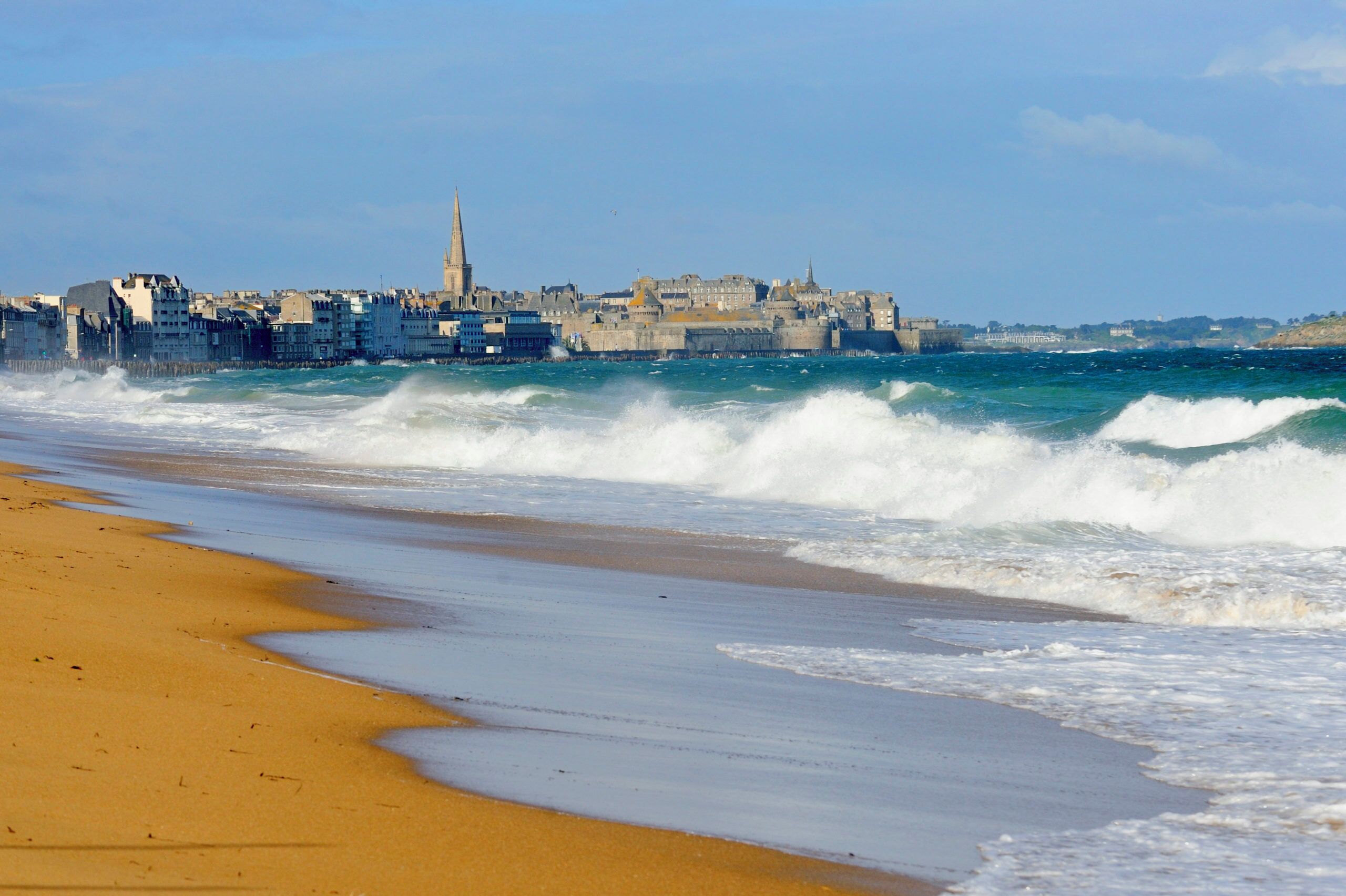 Un paisaje urbano costero con una imponente aguja de iglesia, playa de arena y olas rompiendo en la orilla.