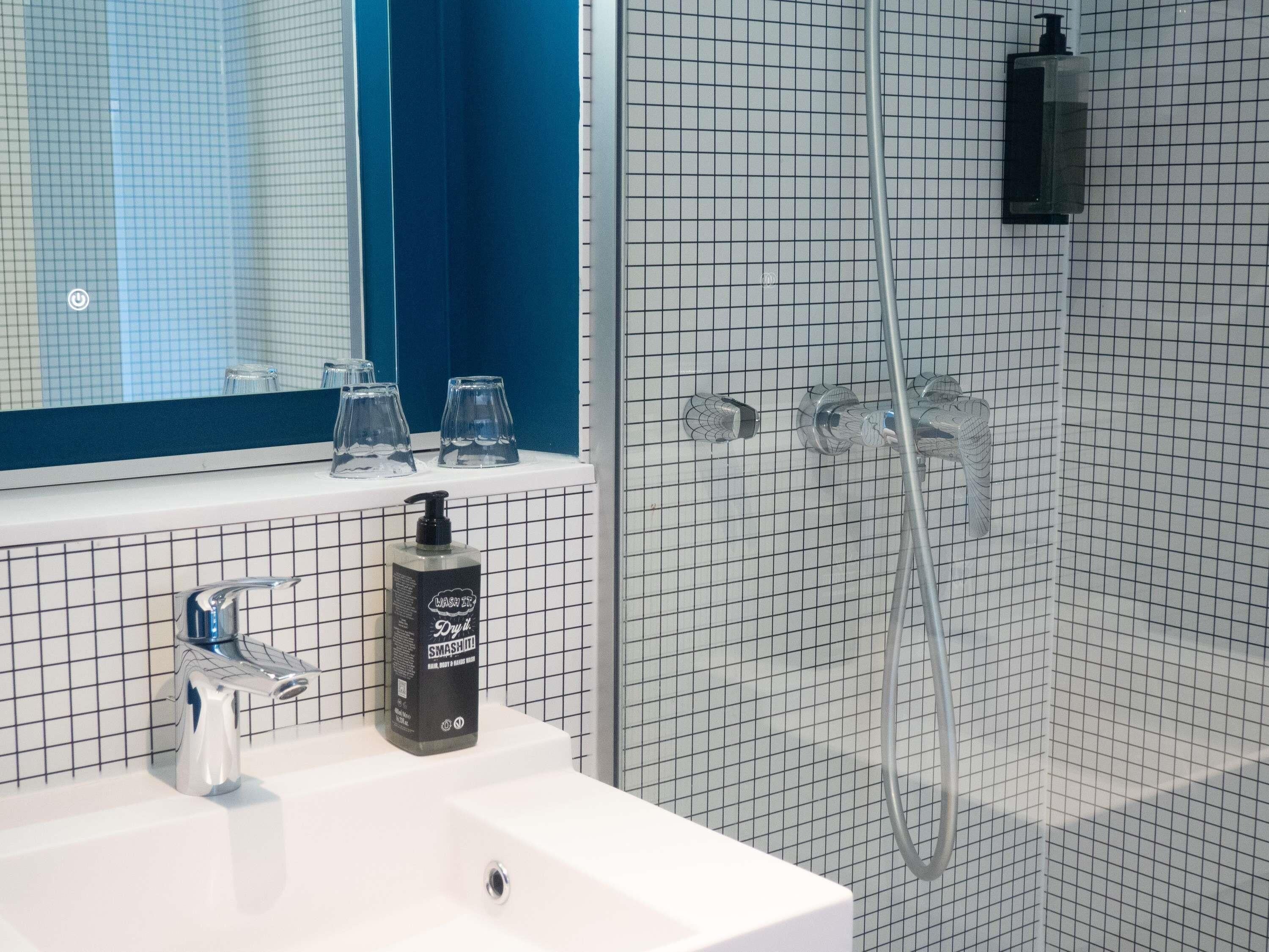 Un baño con un lavamanos blanco, un espejo, un cabezal de ducha manual y una botella de jabón.