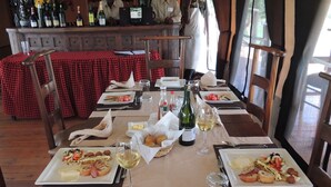 A dining area with a long table set for a meal, a bar in the background, and a view of the outdoors. - Loyk Mara Camp (Maasai Mara)