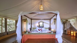 A four-poster canopy bed with a wooden chest in front, a bedside table with a lamp, and a view of the outdoors through large windows. - Loyk Mara Camp (Maasai Mara)