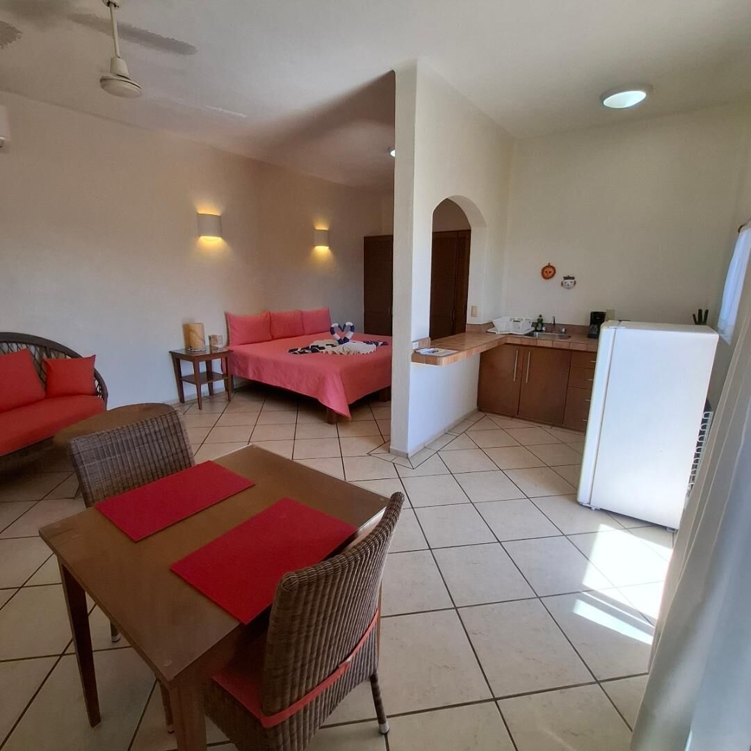 Habitación de hotel compacta con cama, una pequeña mesa de comedor con sillas, kitchenette y ventilador de techo.