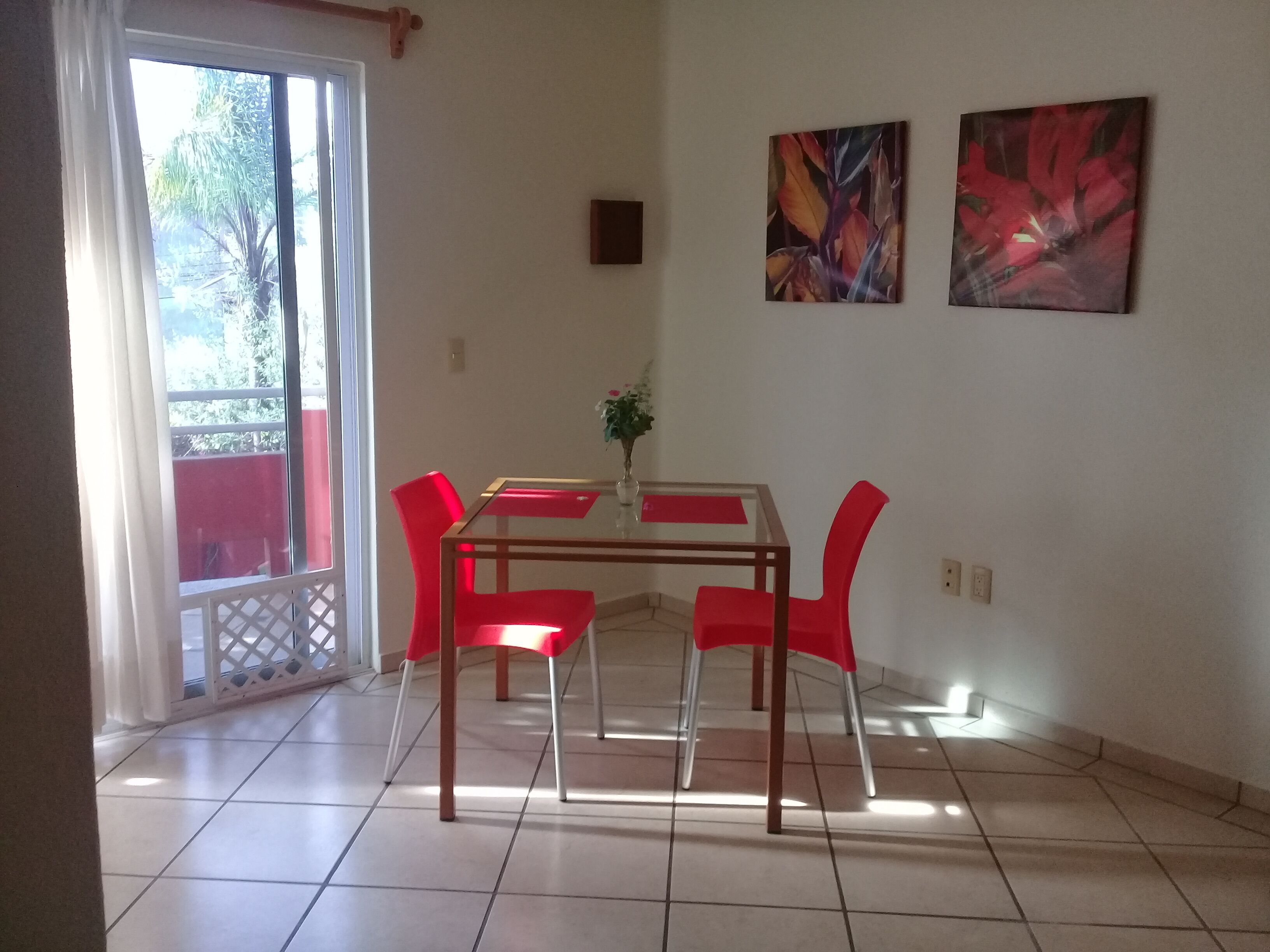 Un área de comedor con una mesa de vidrio y sillas rojas, una planta en maceta y dos obras de arte en la pared.