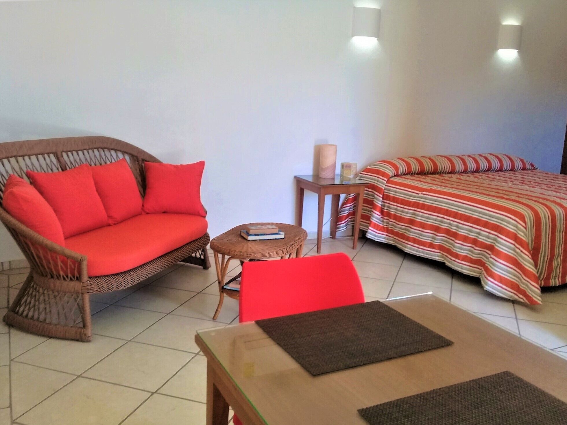 Una habitación con un sofá de mimbre, una silla roja, una mesita con un libro, una cama con una colcha a rayas y una mesita de noche con una lámpara.
