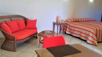 Una habitación con un sofá de mimbre, una silla roja, una mesita con un libro, una cama con una colcha a rayas y una mesita de noche con una lámpara.