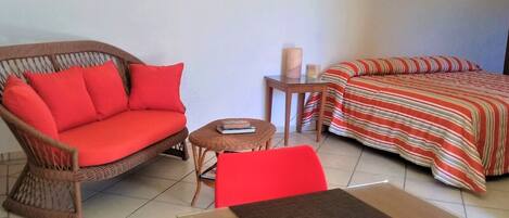 Una habitación con un sofá de mimbre, una silla roja, una mesita con un libro, una cama con una colcha a rayas y una mesita de noche con una lámpara.