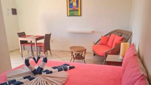 A hotel room with a large bed, a dining area with a table and chairs, a wicker chair, and a painting on the wall.