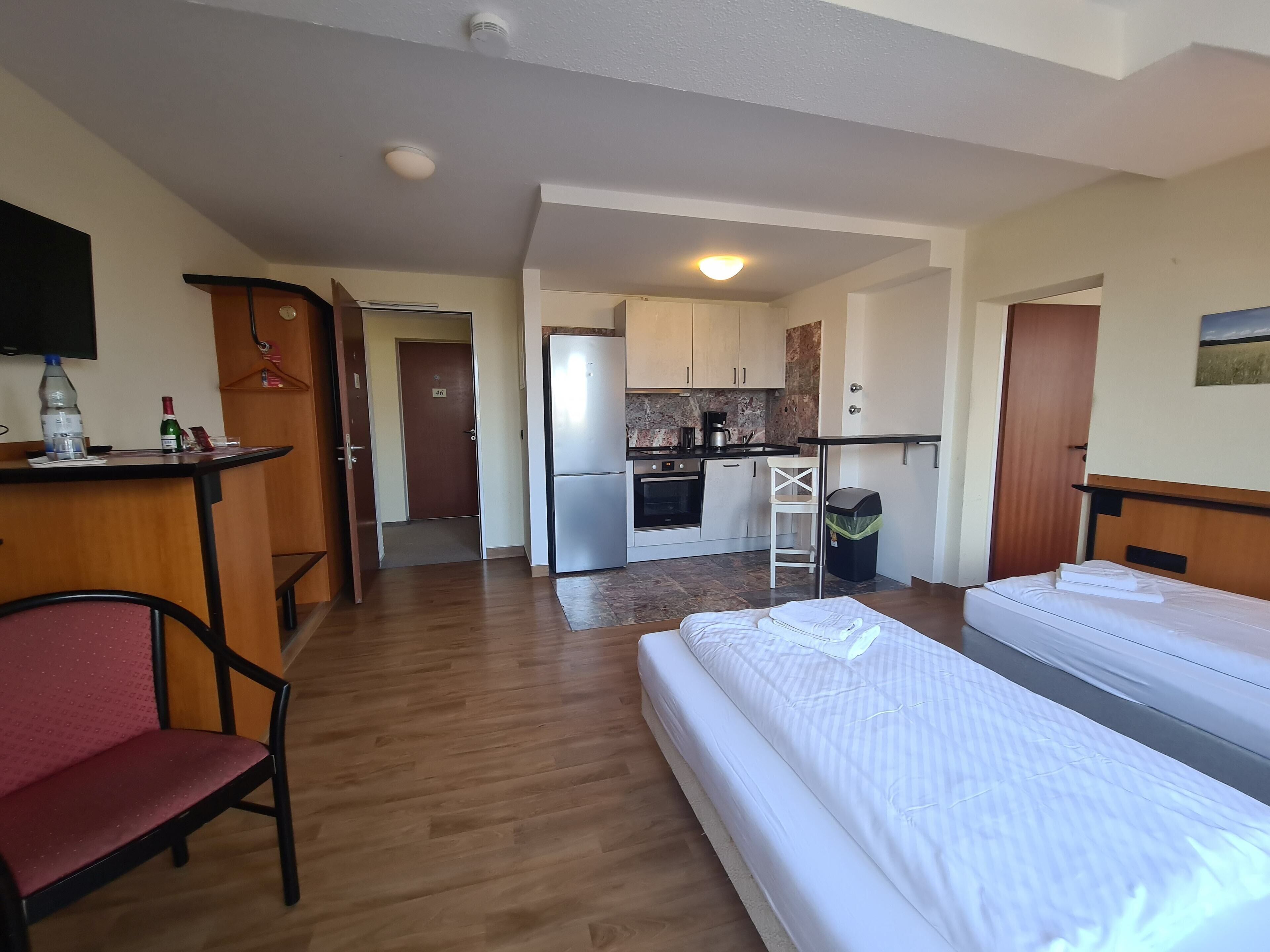 Quarto de hotel compacto com cozinha compacta, uma cama de solteiro, uma pequena mesa de trabalho com cadeira e televisão.