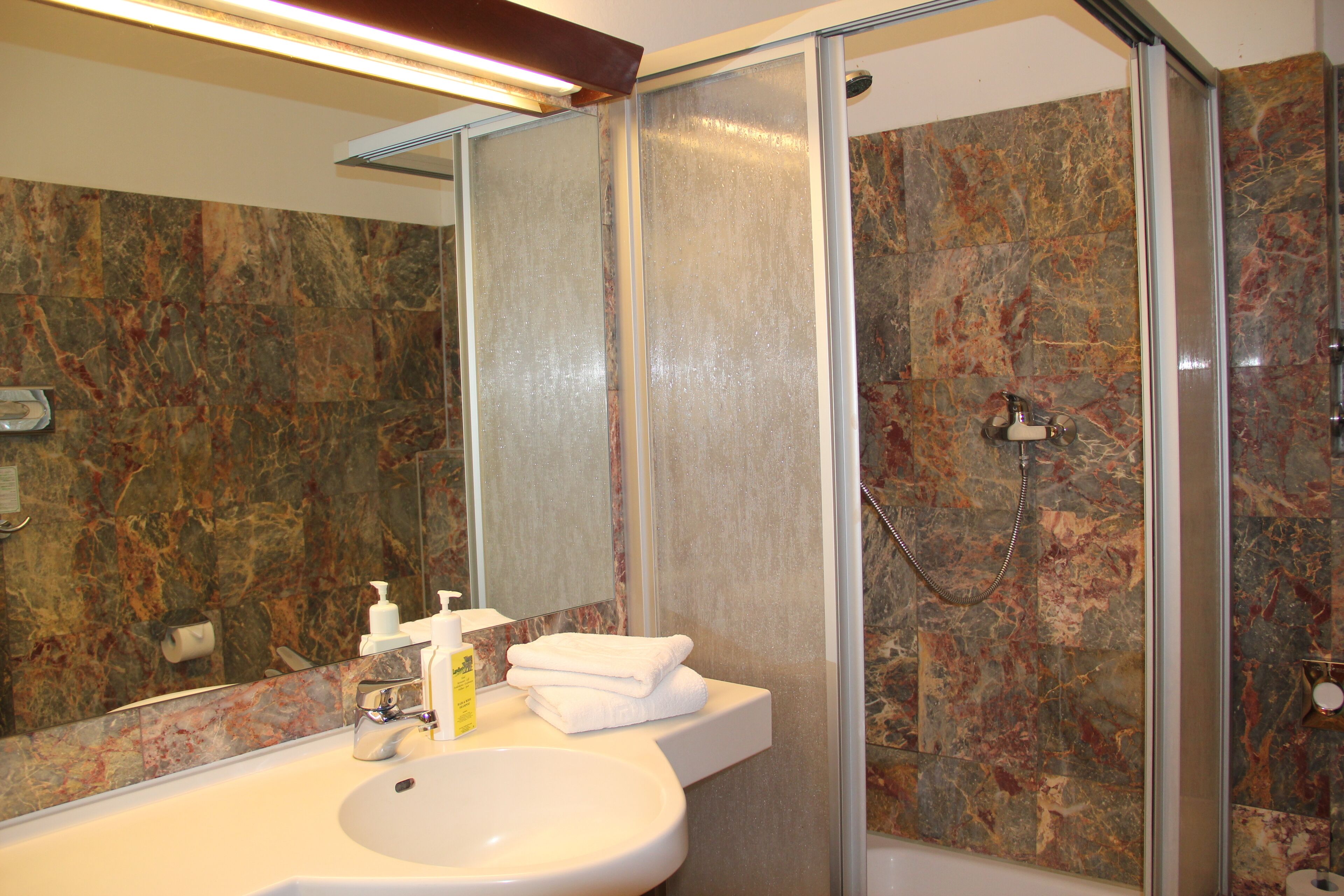 A bathroom with a marble wall, a sink with toiletries, and a glass-enclosed shower.