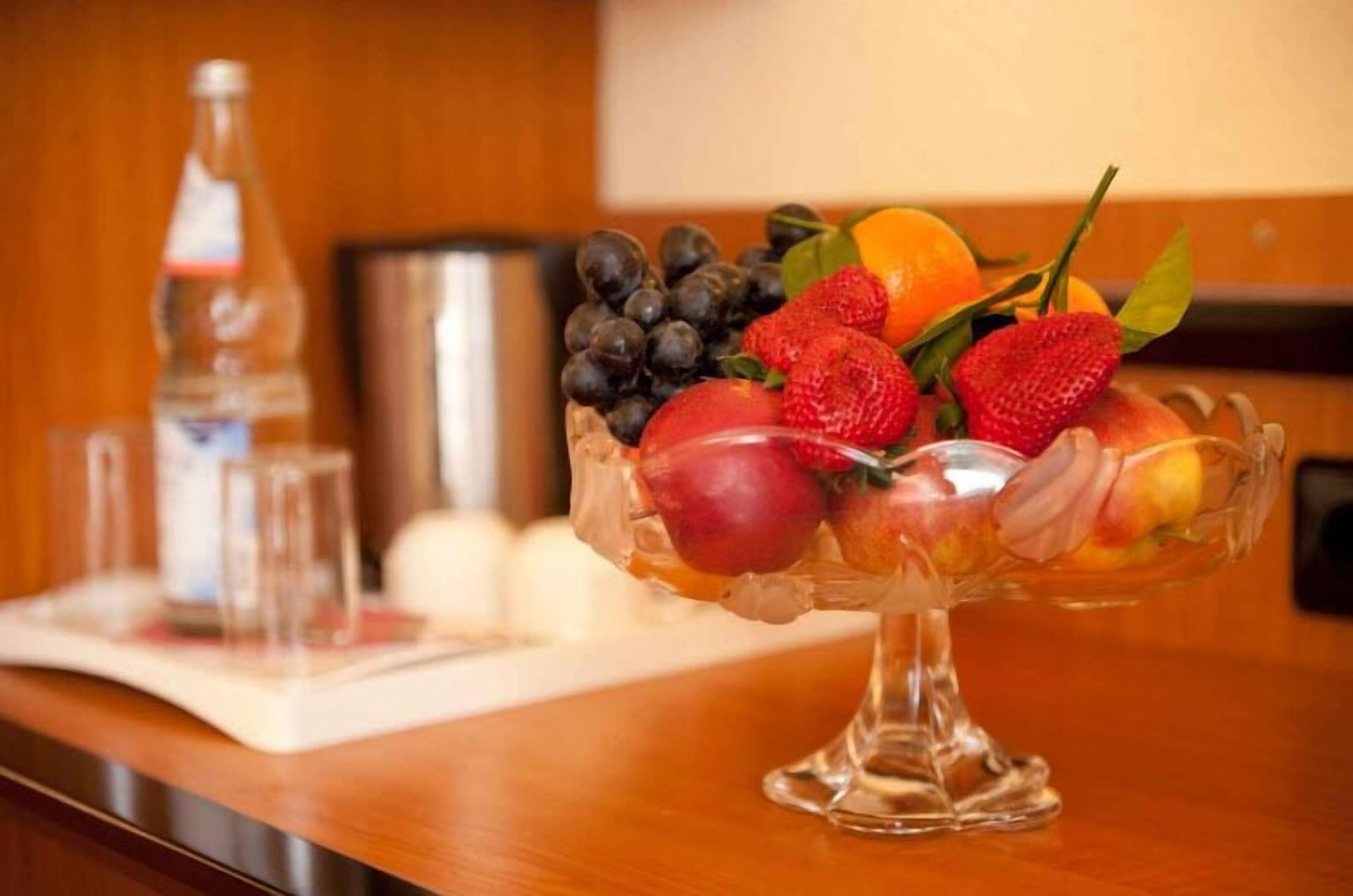 Un frutero sobre una mesa de madera, con una botella y copas al fondo.