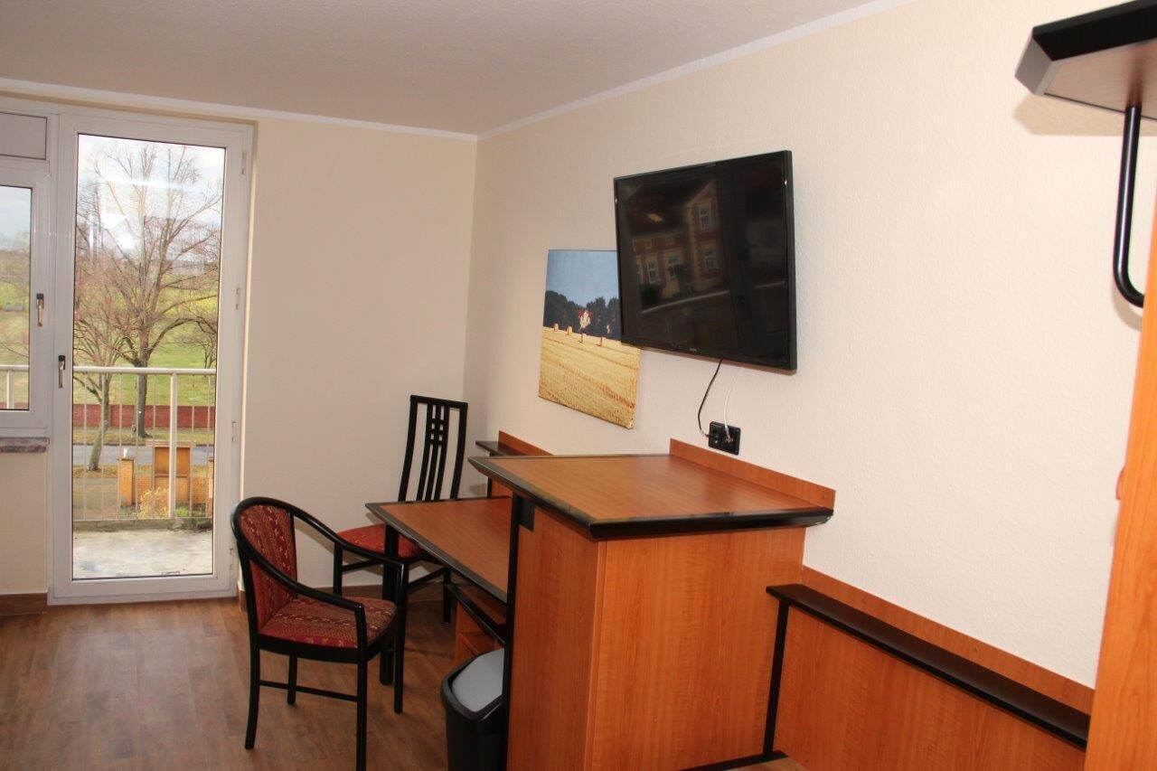 A room with a wooden desk, two chairs, a television mounted on the wall, and a door leading to a balcony.