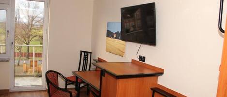 A room with a wooden desk, two chairs, a television mounted on the wall, and a door leading to a balcony.