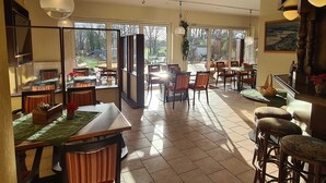 A spacious dining area with multiple tables and chairs, large windows offering a view of the outdoors, and a mix of hanging and pendant lighting. - Landhotel Löwenbruch (Ludwigsfelde)