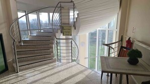 A hotel lobby with a staircase leading to upper floors, a sign indicating room numbers, and a view of the outdoors. - Landhotel Löwenbruch (Ludwigsfelde)