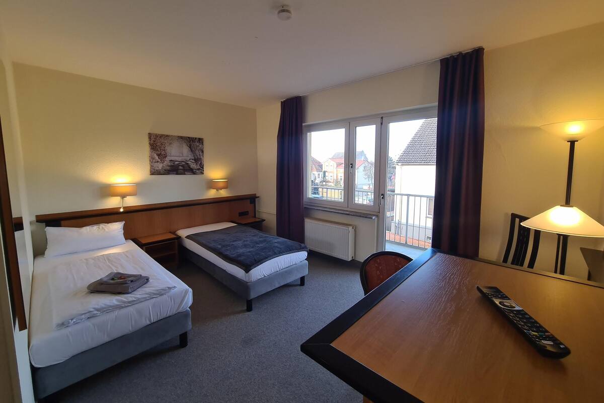 A hotel room with two single beds, a wooden desk, a chair, a table lamp, and a view of buildings through the window.