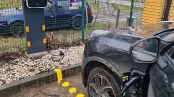 Un espace de stationnement équipé de bornes de recharge pour véhicules électriques, une clôture et un bâtiment en arrière-plan.