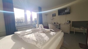 A hotel room with a large bed, a desk, a chair, a TV, and a window with a view of trees. - Landhotel Löwenbruch (Ludwigsfelde)