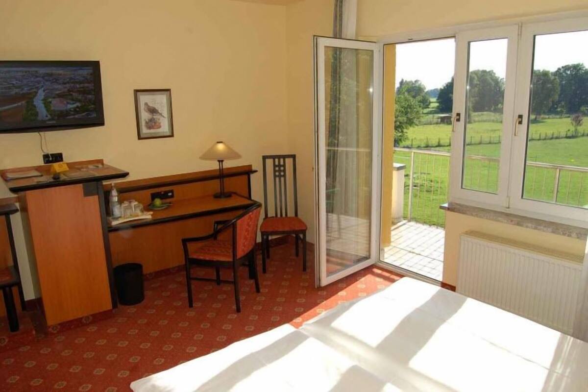 A room with a desk, chair, and a television on the wall. There is a sliding glass door leading to an outdoor area with a view of greenery.