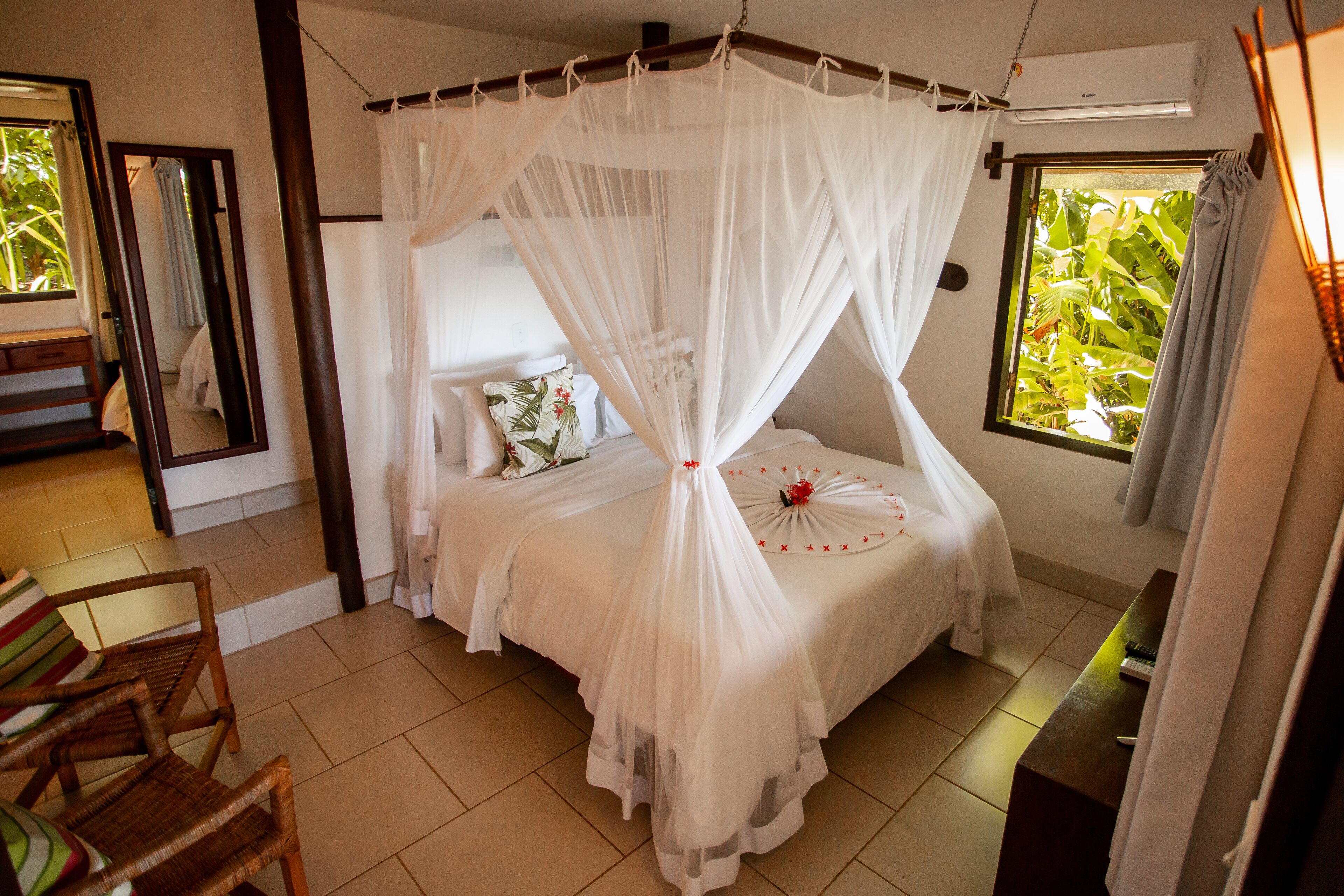 A bedroom with a four-poster bed, a sitting area with chairs, and a large window with a view of greenery.
