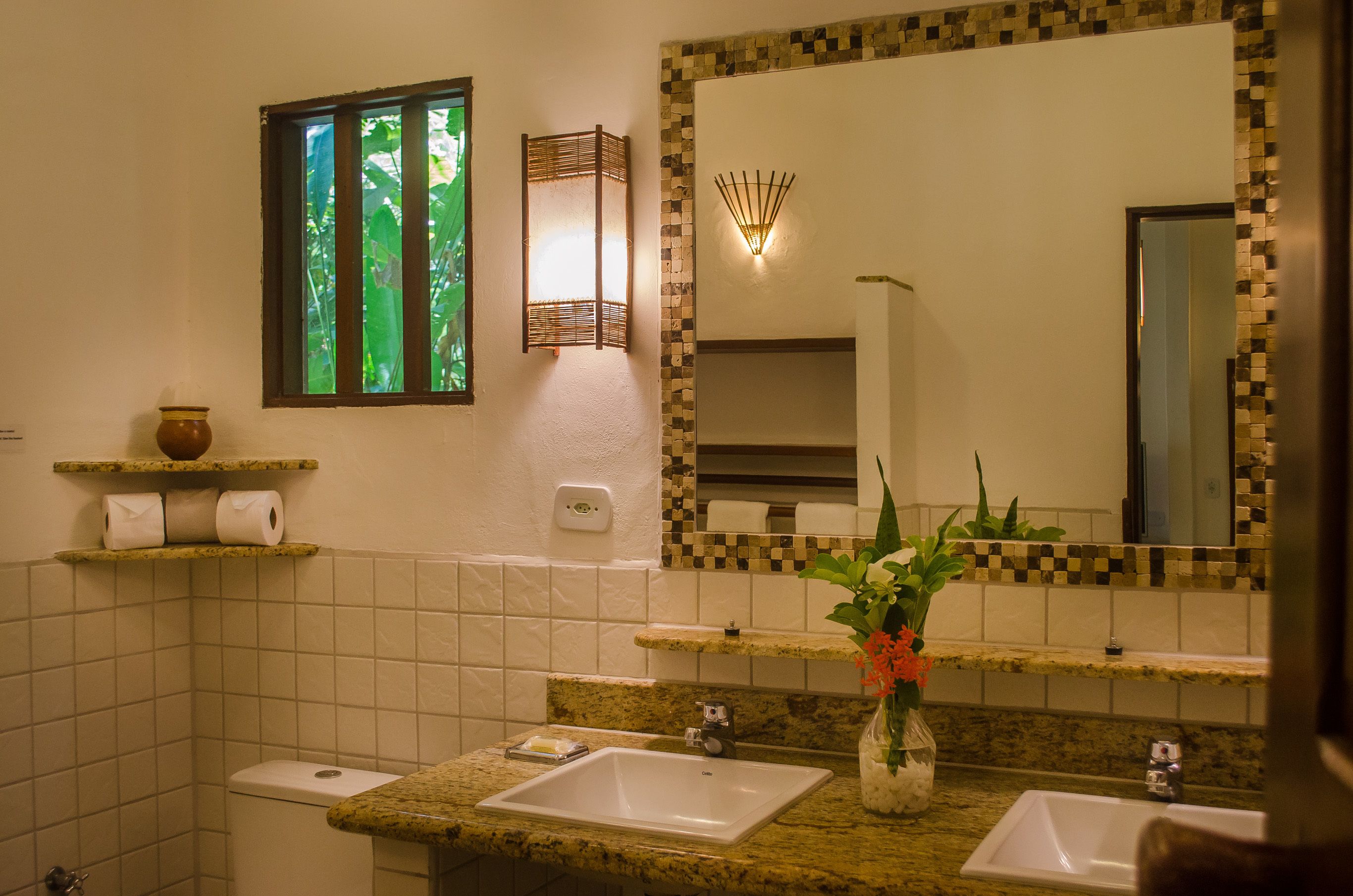 Een badkamer met een toilet, twee wasbakken, een spiegel, een plank met toiletartikelen en een raam met uitzicht op groen.