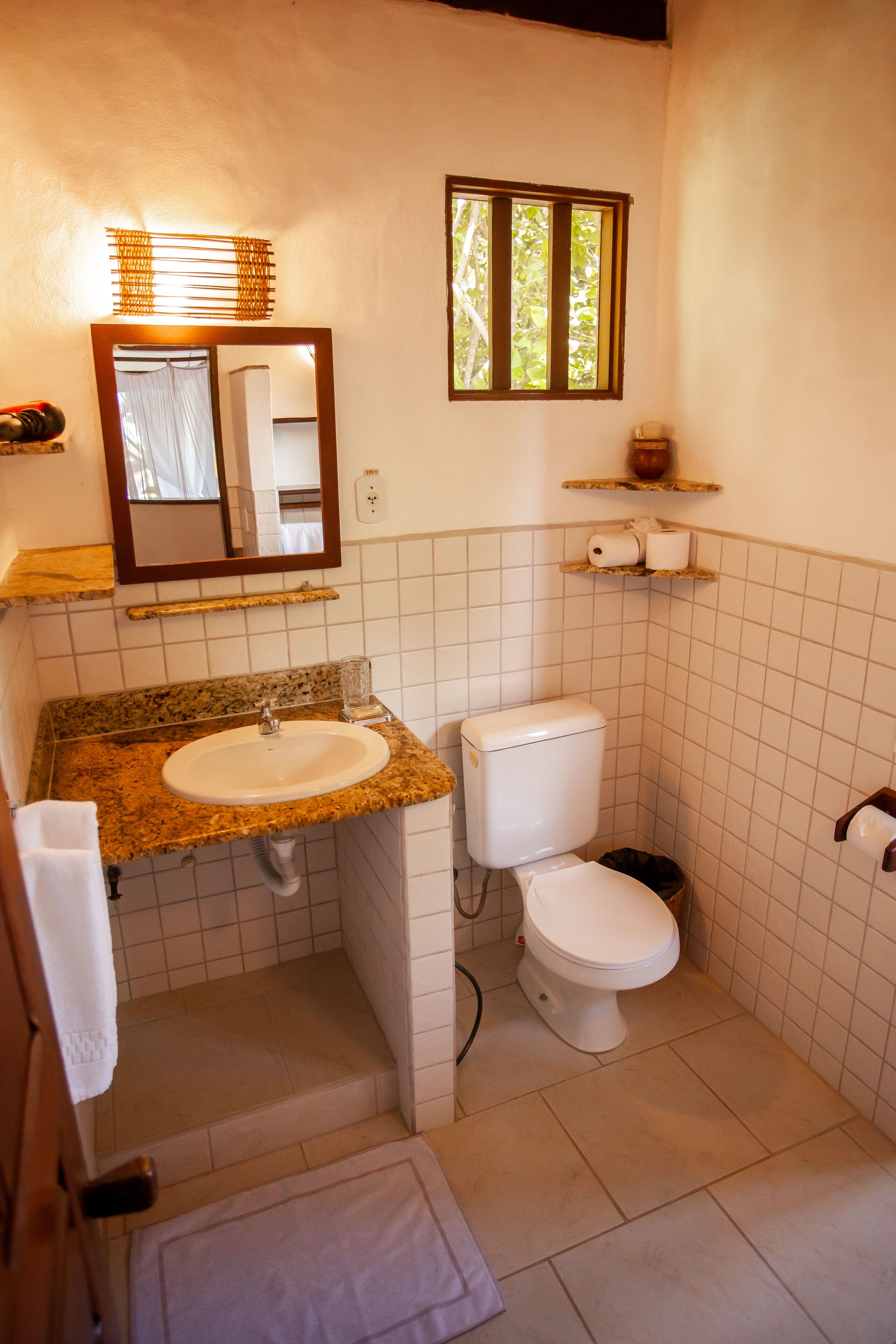 A bathroom with a granite countertop sink, a toilet, and a mirror above the sink. There are shelves above the toilet and a window above the mirror.