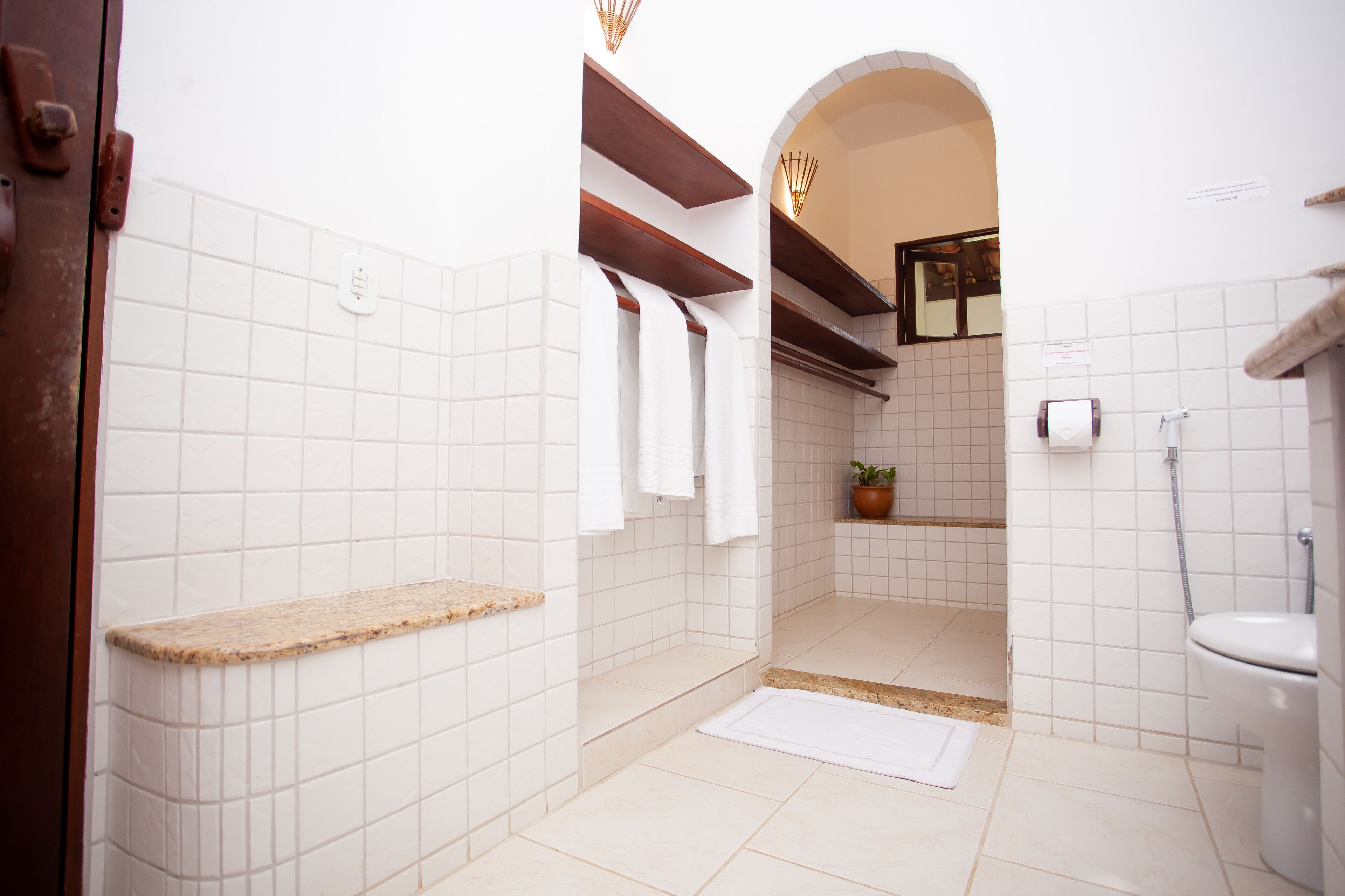 Une salle de bain avec des carreaux blancs, une étagère en bois, un espace douche et des toilettes.