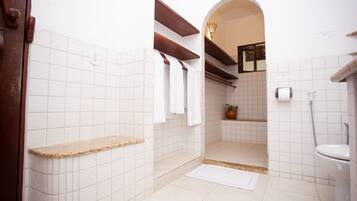 A bathroom with white tiles, a wooden shelf, a shower area, and a toilet.