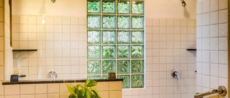 A bathroom with a shower, a sink with a potted plant, and a window with green foliage.