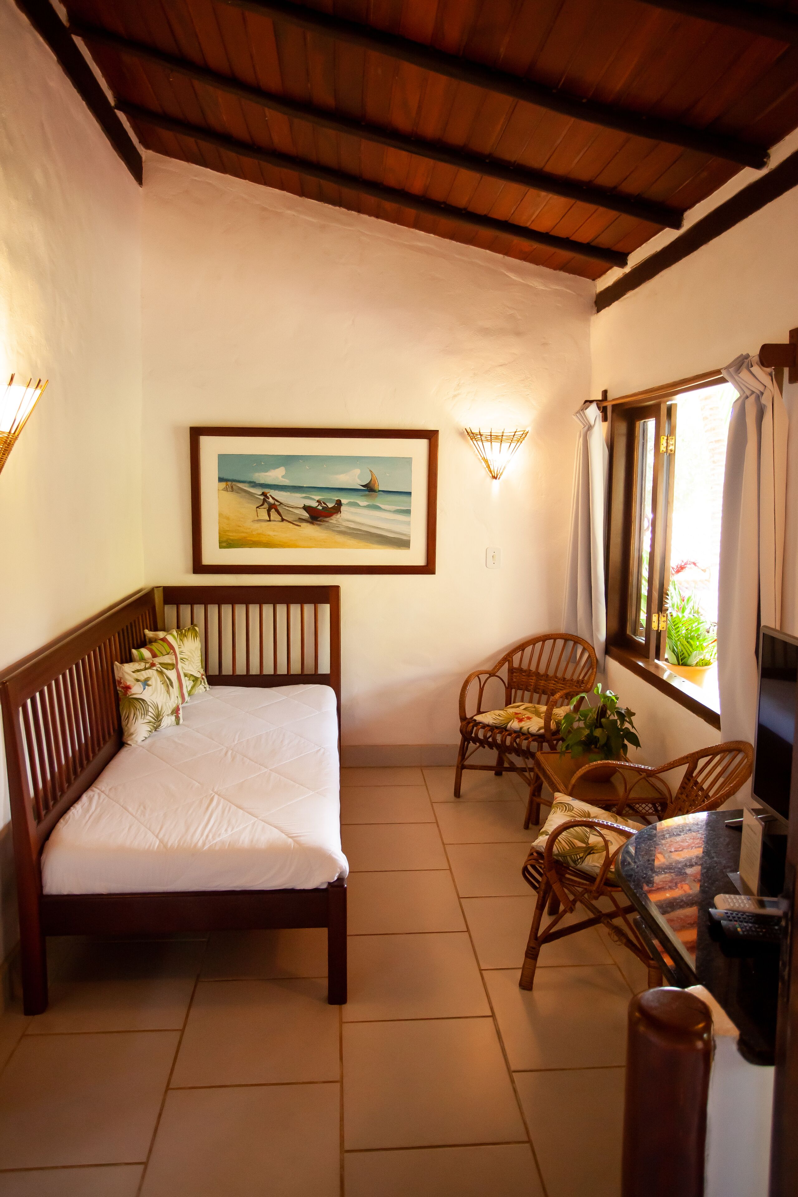 A bedroom with a bed, two wicker chairs, a small table, a painting on the wall, and a window with curtains.