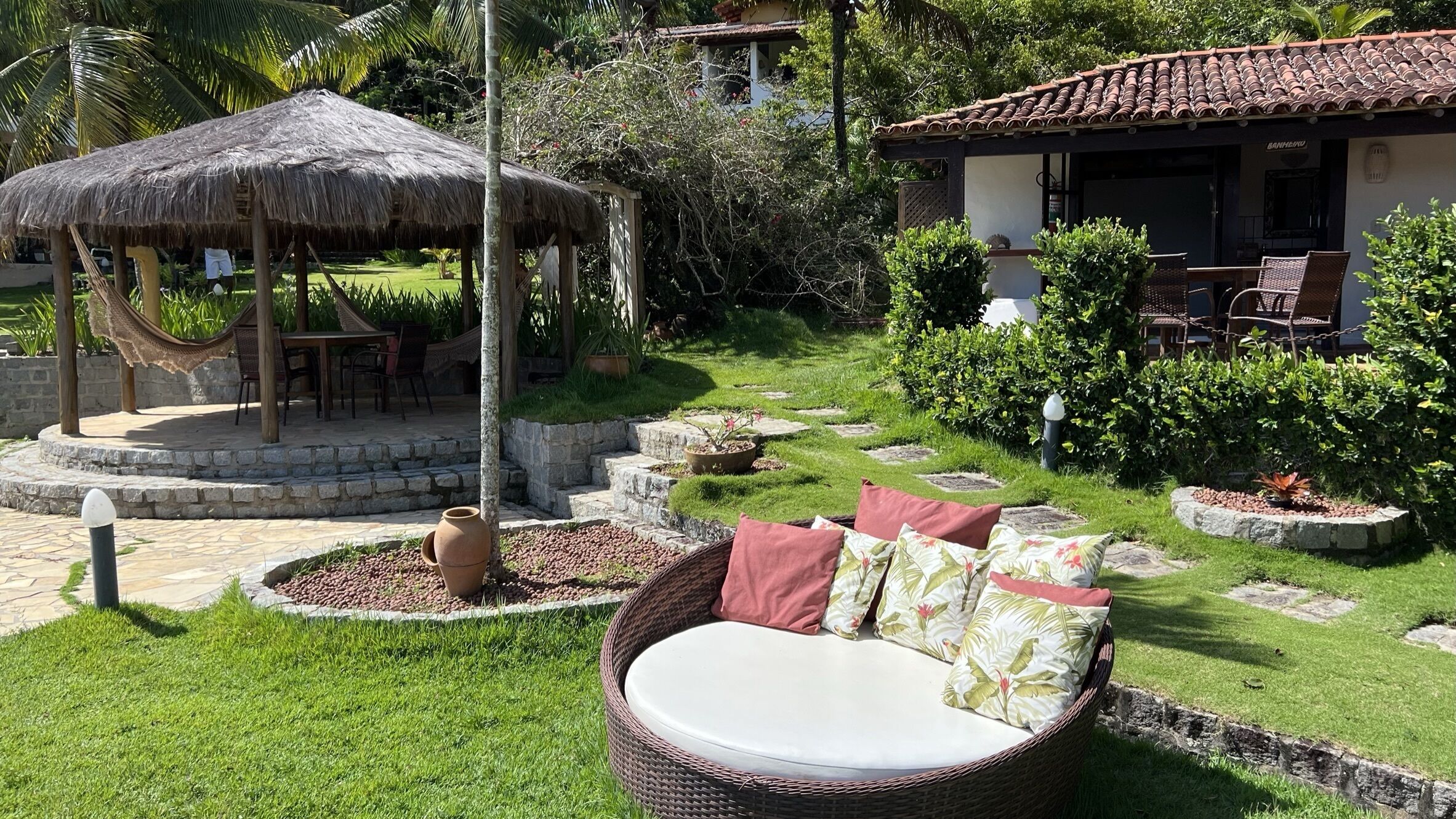 A garden with a wicker sofa, a thatched-roof structure, and a building with a tiled roof.