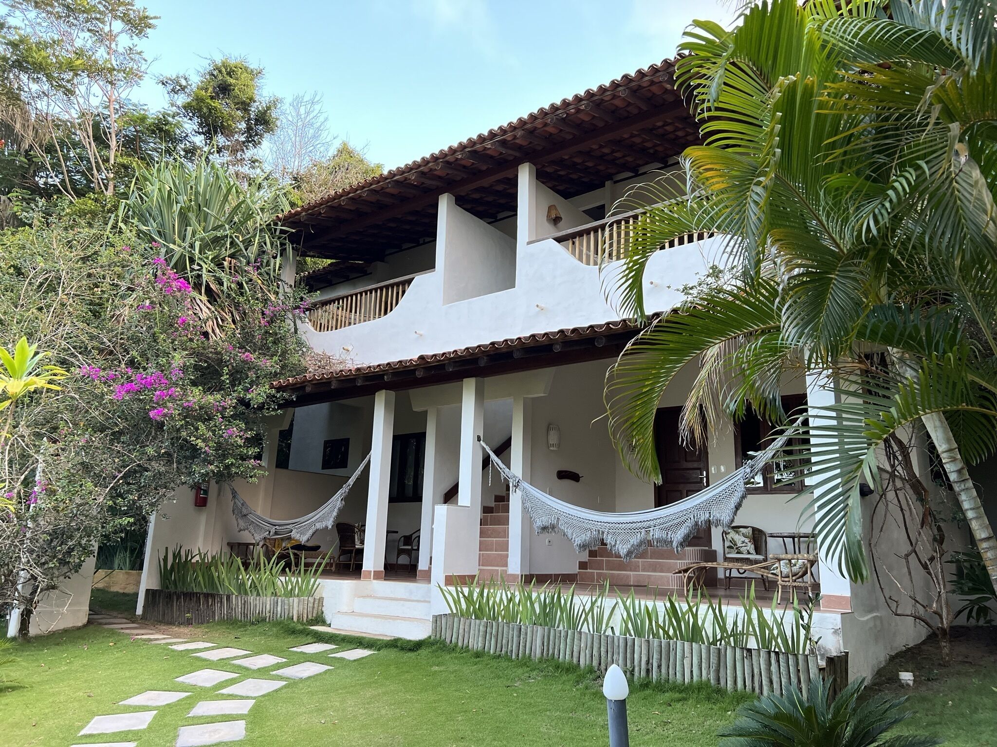 Una casa de dos pisos con un porche cubierto, una hamaca y un jardín con plantas tropicales.