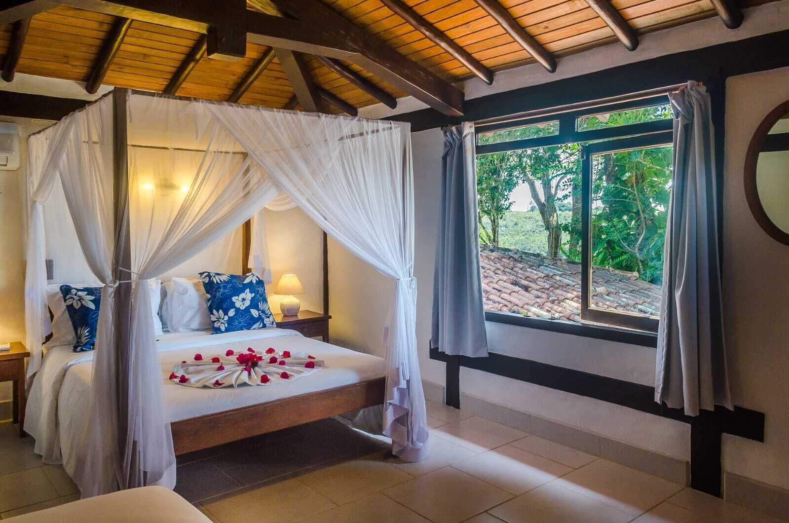 A four-poster bed with a canopy, a bedside table with a lamp, and a view of greenery through a window.