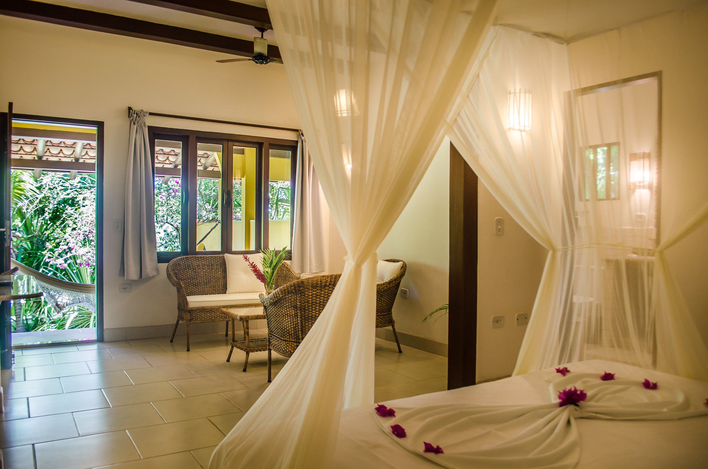 A room with a bed covered in white sheets and a canopy, two wicker chairs, a small table, and a view of greenery outside.