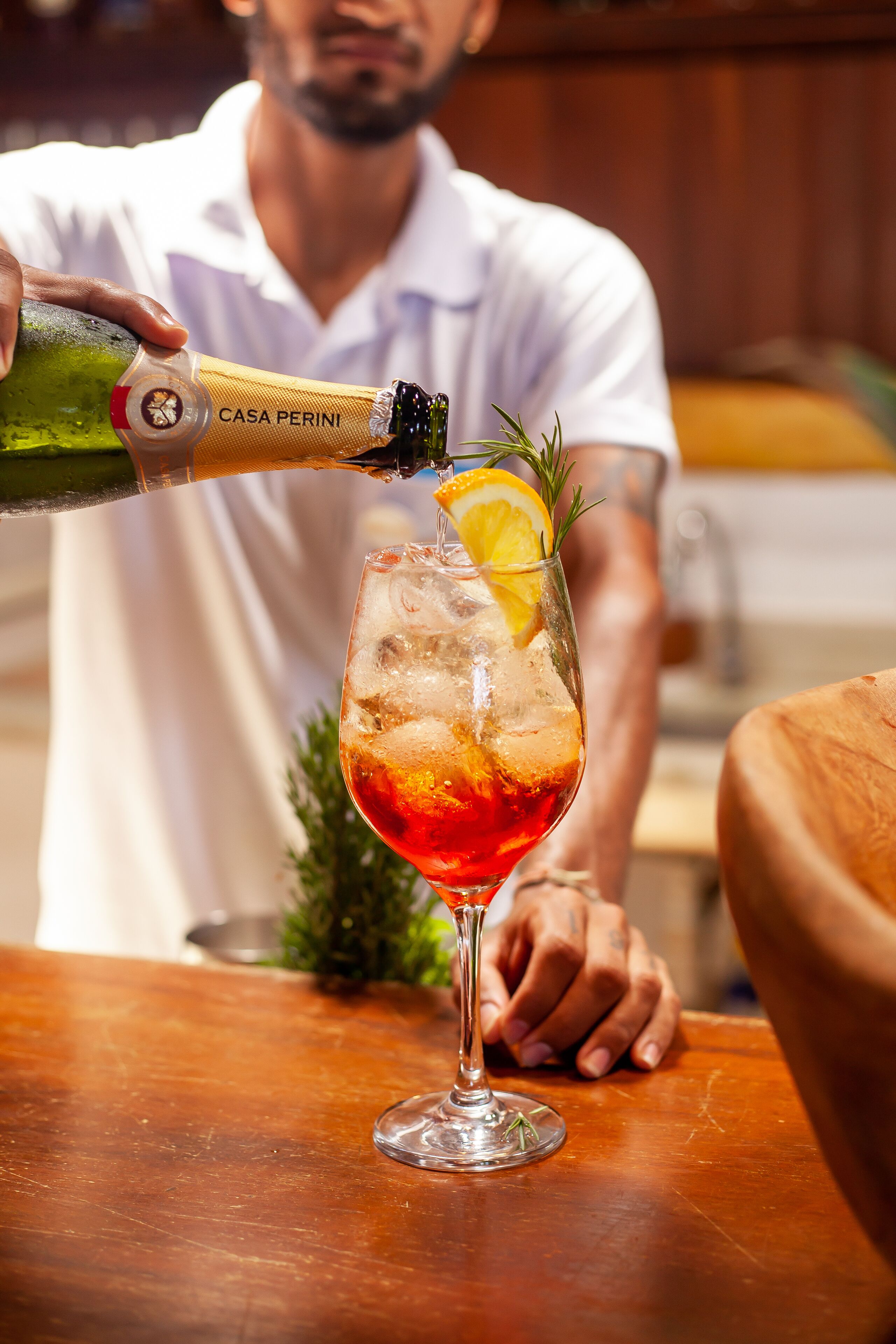 A person pouring a drink from a bottle labeled "CASA PERINI" into a glass with ice, garnished with a slice of orange and a sprig of rosemary.